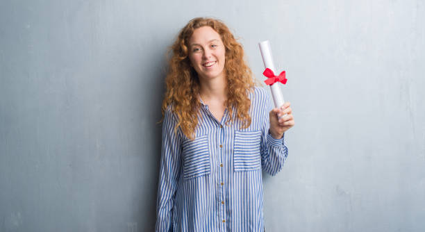 femme d’affaires jeune rousse au fil du mur gris grunge comprenant diplôme avec un visage heureux, debout et souriant avec un sourire confiant, montrant les dents - women business graduation casual photos et images de collection