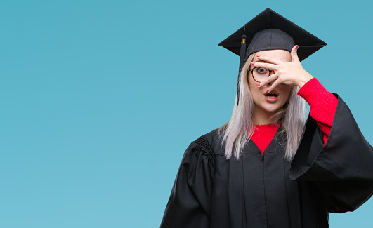 Young blonde woman wearing graduate uniform over isolated background peeking in shock covering face and eyes with hand, looking through fingers with embarrassed expression.