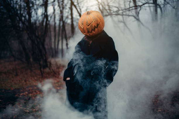A man in a long black coat with a pumpkin instead of a head in a terrible night forest, damp and fog. Happy Halloween story concept