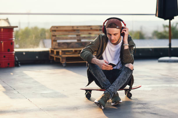 istock Young handsome man listening music in headphones and using smartphone while sitting on skateboard on the rooftop of industrial building 1050929898
