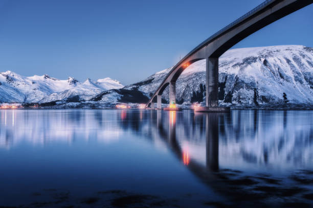 Bridge with illumination, snow covered mountains, village and blue sky with reflection in water. Night landscape with bridge, snowy rocks reflected in sea. Winter in Lofoten islands, Norway. Roadway