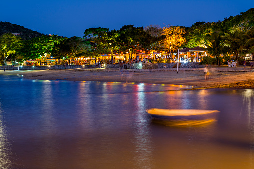 Porto da Barra area at Manguinhos beach in Búzios, Rio de Janeiro, Brazil. Crowded restaurants seen in a long exposure night photograph with a fishing boat on the foreground.