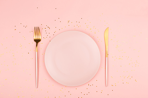 Beautiful pink plate and cutlery on gold confetti and pastel color background. Holiday, Christmas or New Year dinner concept. Flat-lay, top view.