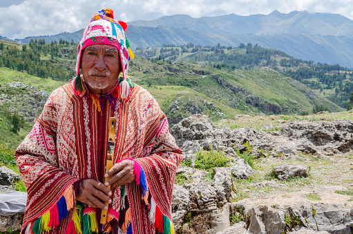 Cusco, Peru; January 24, 2017: Old man Quechua dressed in a colored poncho and a cap of Chullo, plays on the musical instrument quena with a view of the mountains in the background
