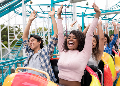 Happy group of people having fun in an amusement park riding on a rollercoaster with arms up and screaming - lifestyle concepts