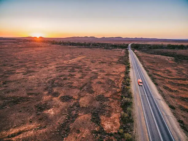 Photo of Red car driving on rural road passing through Australian outback leading to Flinders Ranges peaks at sunset
