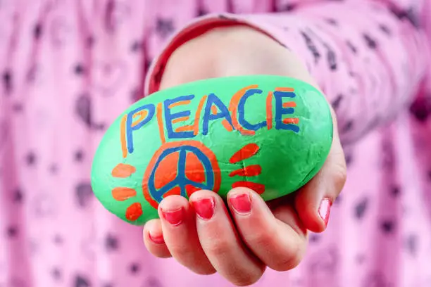Photo of Child holding painted rock wit PEACE lettering and symbol. Focus on foreground