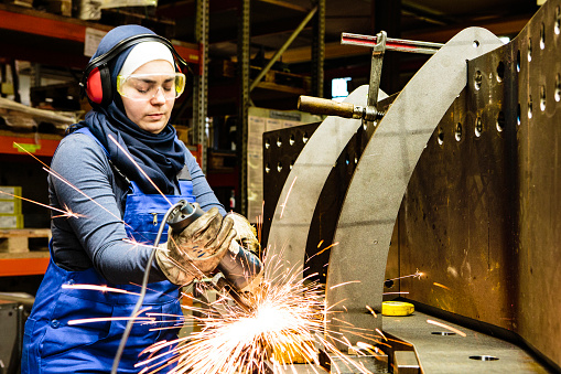Young female trainee welds steel with grinder in workshop, flying sparks