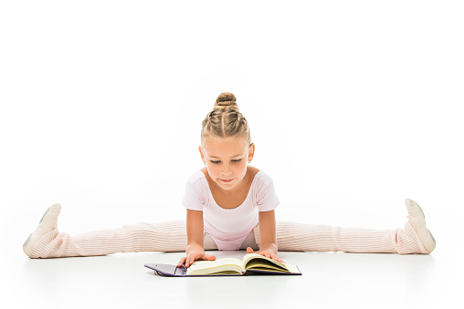 concentrated little ballerina reading book and doing twine isolated on white background