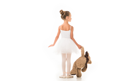 rear view of little ballerina in tutu practicing with teddy bear isolated on white background