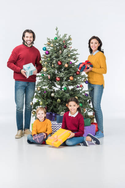 istock Happy parents with gift boxes standing while siblings sitting near christmas tree isolated on white 1051107258