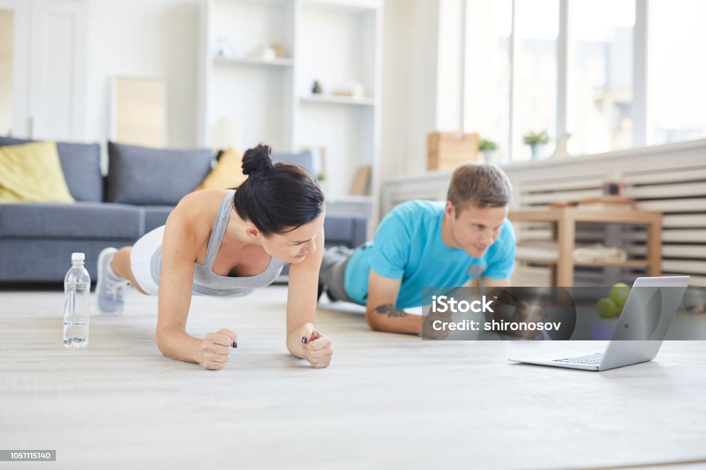 Repeating after online trainer Contemporary young active couple in sportswear doing planks on the floor in front of laptop while watching online workout Exercising Stock Photo