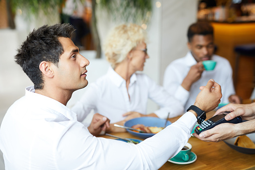 Serious handsome young manager in white shirt sitting at table with friends and paying for lunch with smartwatch in restaurant