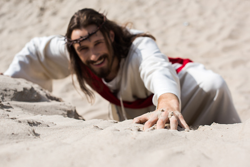 high angle view of smiling Jesus in robe, red sash and crown of thorns climbing sandy hill in desert