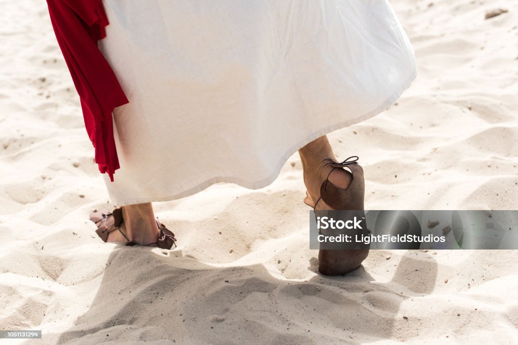 cropped image of Jesus in robe, sandals and red sash walking on sand in desert Jesus Christ Stock Photo