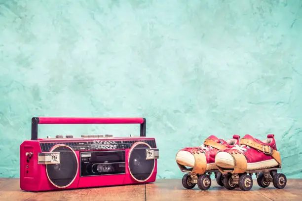 Retro designed radio cassette tape recorder and classic steel children's roller skates pair with red leather sneakers front aquamarine concrete wall textured background. Vintage style filtered photo