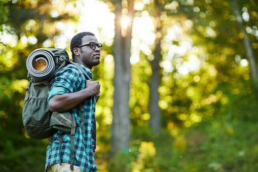 African-american guy carrying backpack on his back while moving in the forest on summer weekend