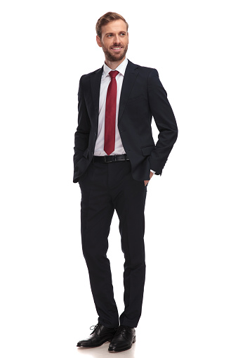 relaxed businessman smiles and looks to side while standing on white background with hands in pockets