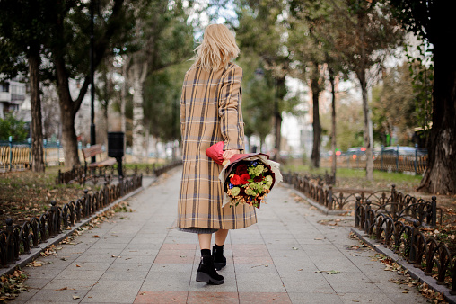 Back view of blonde woman in plaid coat walking with a bouquet of flowers in the park