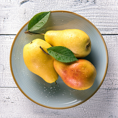 Square image of wet ripe golden yellow pears and green leaves on round blue plate on old white wooden table surface. Selective focus. View from above. Healthy eating concept.