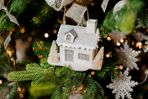 Stylish Christmas tree decorative toy in shape of white house covered with snow hanging on green branch near snowflakes