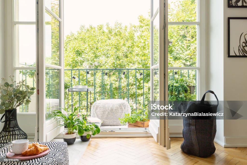 Open glass door from a living room interior into a city garden on a sunny balcony with green plants and comfy furniture Balcony Stock Photo