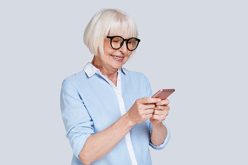 Beautiful senior woman using her smart phone and smiling while standing against grey background
