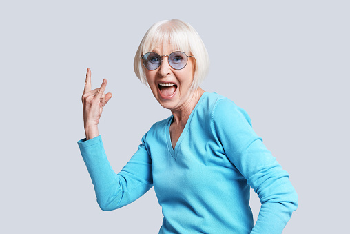 Playful senior woman making rock on sign and smiling while standing against grey background