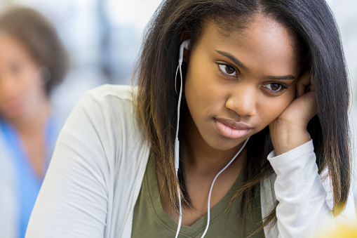 In this closeup, a teenage girl sits with her head resting on her hand.  She wears a frustrated expression as she listens to music on her earbuds.  Her mother can be seen in the background trying to get her attention.