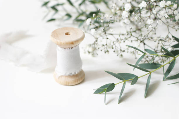 feminine floral scene. close-up of spool of silk ribbon, eucalyptus parvifolia leaves and babys breath gypsophyla flowers on a white table background. wedding styled stock photography. selective focus - table wedding flower bow imagens e fotografias de stock