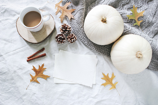 Autumn breakfast in bed composition. Blank cards mockup. Cup of coffee, white pumpkins, sweater, oak leaves and pine cones on linen background. Thanksgiving, Halloween. Flat lay, top view.