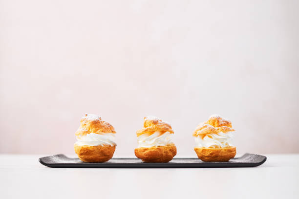 Delicious profiteroles with cream and powdered sugar on a plate. Delicious cream puff cakes with cream and powdered sugar on a black plate on white marble table over pink background. Selective focus, copy space. cream cake stock pictures, royalty-free photos & images