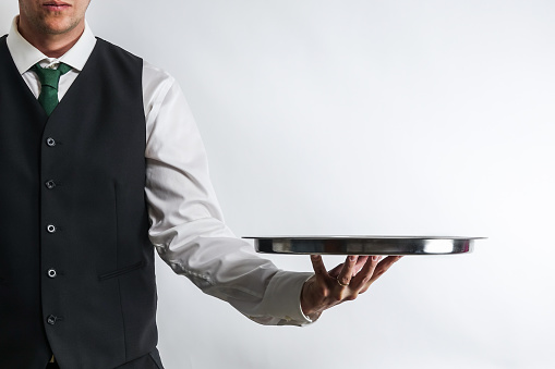 Butler / waiter in white shirt and black suit vest carrying an empty silver tray. Copy space for text on white background.