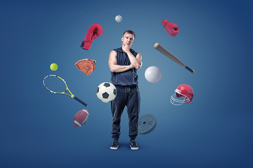 A thoughtful muscular man stands surrounded by different sport gear flying around him. Individual or group sport. Choosing your activity type. Staying fit with sport.