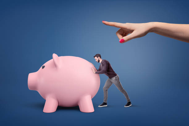 una mano mujer gigante con un dedo apuntando en algún lugar y un hombre empujando un banco grande razonable en esa dirección. - piggy bank savings pig currency fotografías e imágenes de stock