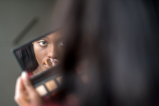 Portrait of Young black woman looking at the make up mirror while fixing her make up.