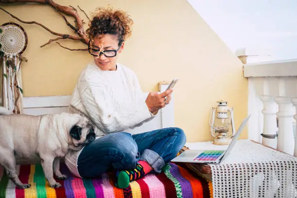 Happy alternative young cheerful woman working at home with a new business way. sitting on a bench outdoor at home with best friend old pug together. colorful bench and lifestyle. laptop and phone to communicate and internet
