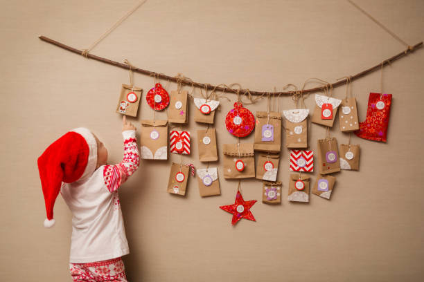 kind schaut der adventskalender. babymädchen in eine weihnachtsmütze und pyjamas zeigt auf ersten geschenk - adventskalender stock-fotos und bilder