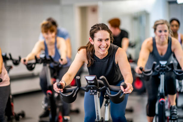 Women Working Out A group of adults are indoors in a fitness center. They are riding exercise bikes. One woman is smiling in the foreground. exercise class stock pictures, royalty-free photos & images