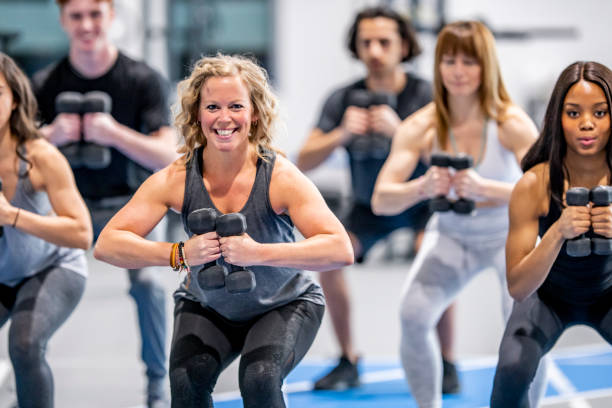 Doing Exercises Together A multi-ethnic group of adults are indoors in a fitness center. They are lifting dumbbells and squatting while smiling at the camera. gym group class stock pictures, royalty-free photos & images