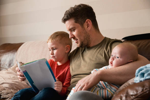 Father and Son Reading a Book Mid-adult father is holding his newborn baby boy in his arm and then reading though the book with his son under the other arm. They all all sitting on the sofa in the living room. single father stock pictures, royalty-free photos & images