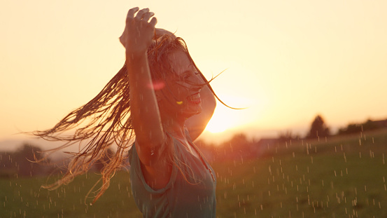 CLOSE UP, LENS FLARE: Attractive blonde haired woman playfully spins in the soothing summer rain. Young Caucasian female dancer dances in the rain on a picturesque spring evening in the countryside.