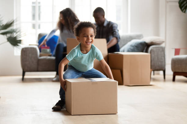 Adorable little boy playing with cardboard box at home Black married couple sitting on couch in living room unpacking boxes with belongings at new home. Little funny African boy carrying playing with cardboard box. New home, relocation and moving concept unpacking photos stock pictures, royalty-free photos & images