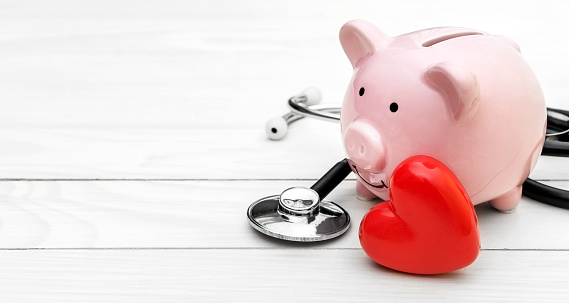 Piggy bank with stethoscope and red heart on white wooden table. Space for text.
