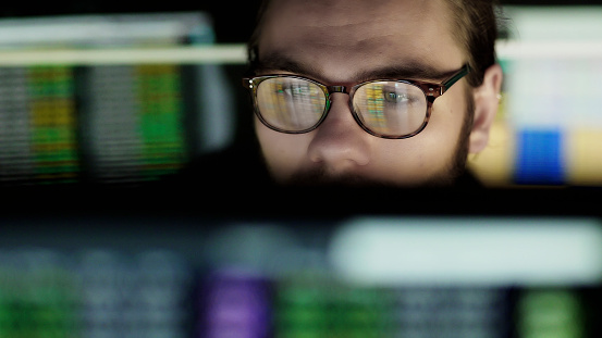 Close up stock image of a young man seated between computer monitors. He’s surrounded by screens displaying changing data as line s of out of focus numbers, he watches this data intently.