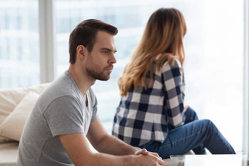 Angry millennial man sit on sofa not looking at beloved woman, lost in thoughts, young couple have disagreement considering breakup or divorce, husband and wife ignore each other close to separation