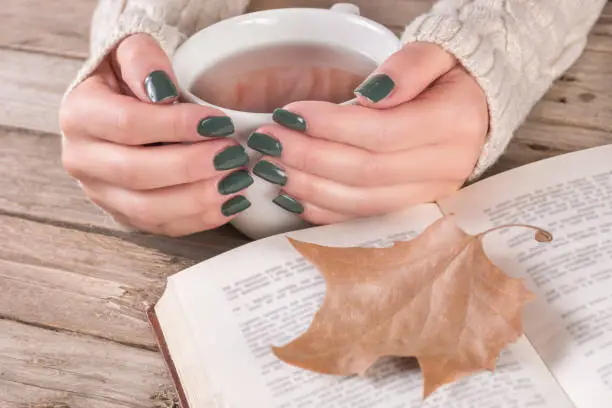 Young girl hands in sweater with olive color manicure holds cup of tea and open book with dry fallen leaf on retro wooden desk. Autumn concept. Close up, selective focus