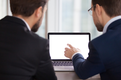 Back view of two male managers or workers look at laptop mock up screen discussing project, colleagues or partners negotiate analyzing statistics and sales working at computer during office meeting