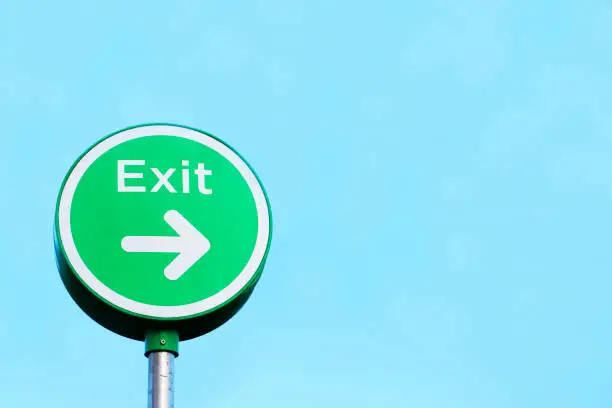 Exit round green sign against blank blue sky with direction arrow in car park uk