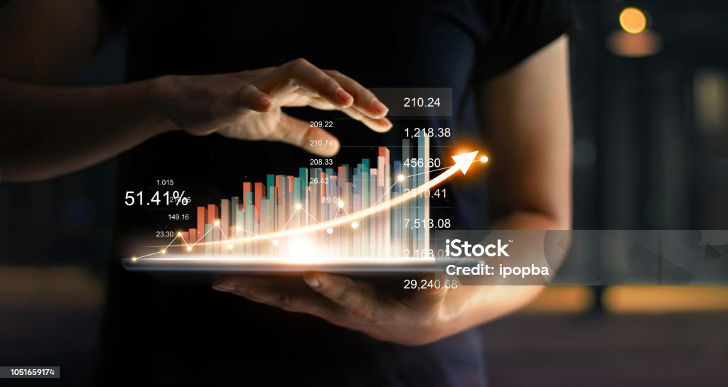Businessman holding tablet and showing a growing virtual hologram of statistics, graph and chart with arrow up on dark background. Stock market. Business growth, planing and strategy concept. Business Stock Photo